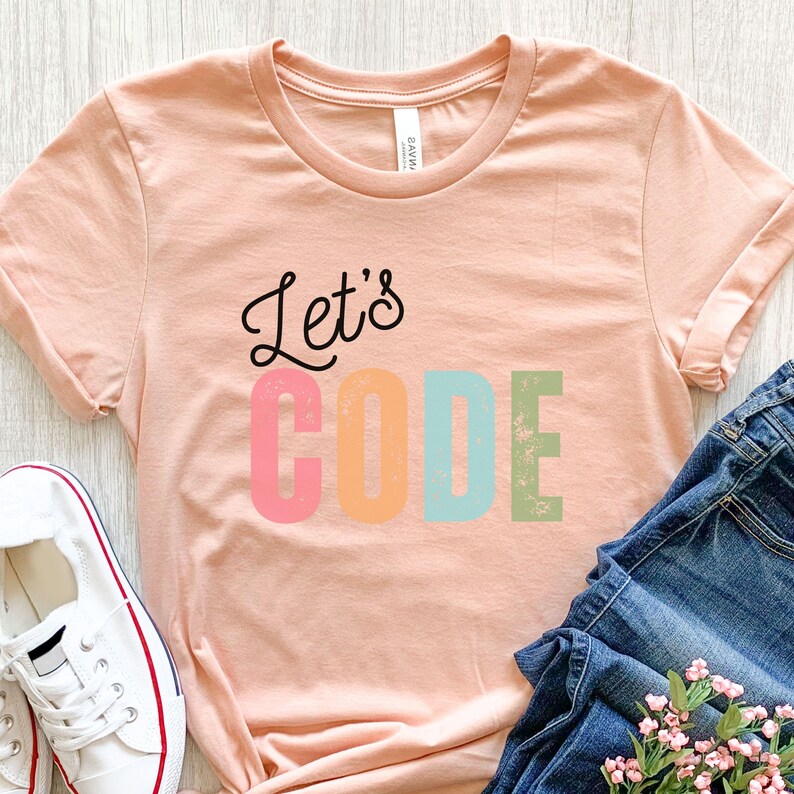 K&ouml;nnte beinhalten: Ein pfirsichfarbenes T-Shirt mit dem Aufdruck "Let's CODE" in Schwarz und Pastellfarben auf der Vorderseite. Das Shirt wird mit einer blauen Jeans und wei&szlig;en Turnschuhen kombiniert.