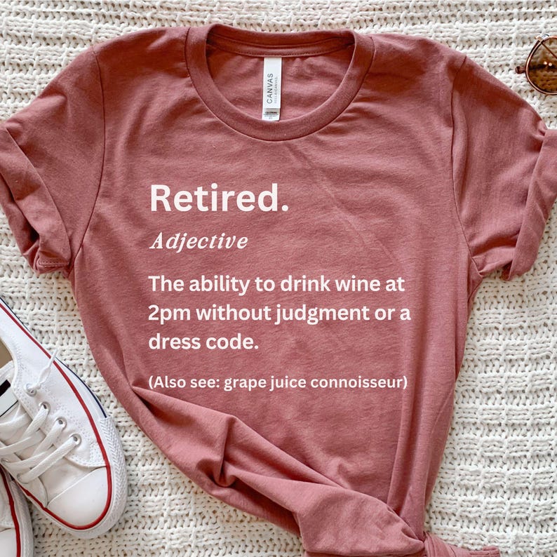 May include: A rose-coloured t-shirt with the word "Retired" in white, followed by a definition. The definition reads: "The ability to drink wine at 2pm without judgement or a dress code."