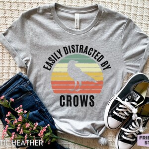 May include: Gray t-shirt with a vintage-style graphic that says "Easily Distracted By Crows" with a silhouette of a crow in front of a sunset.