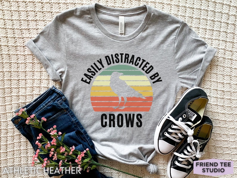 May include: Gray t-shirt with a vintage-style graphic that says "Easily Distracted By Crows" with a silhouette of a crow in front of a sunset.