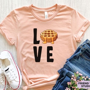 K&ouml;nnte beinhalten: Ein hellrosa T-Shirt mit dem Wort "LOVE" in schwarzer Schrift. Das "O" in LOVE wird durch ein Cartoon-Bild eines Waffels mit Sirup ersetzt.
