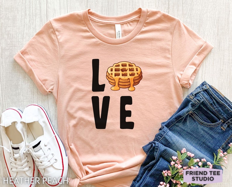 K&ouml;nnte beinhalten: Ein hellrosa T-Shirt mit dem Wort "LOVE" in schwarzer Schrift. Das "O" in LOVE wird durch ein Cartoon-Bild eines Waffels mit Sirup ersetzt.