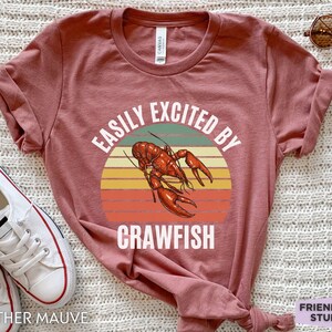 Op de afbeelding: Een heide-paarse t-shirt met een vintage-stijl grafische afbeelding van een rivierkreeft en de tekst "Easily Excited By Crawfish".