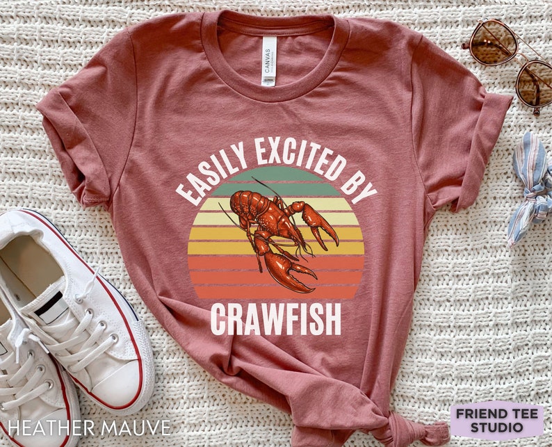 Op de afbeelding: Een heide-paarse t-shirt met een vintage-stijl grafische afbeelding van een rivierkreeft en de tekst "Easily Excited By Crawfish".