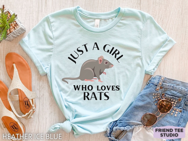 May include: A light blue short-sleeve t-shirt featuring a graphic of a grey rat centered between the black text that reads JUST A GIRL WHO LOVES RATS. The shirt has a crew neckline and is displayed with casual accessories including sandals and denim.