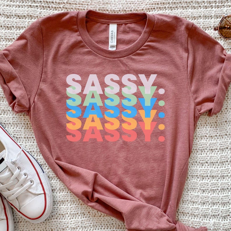 May include: A dusty rose-colored t-shirt with the word "SASSY" stacked five times in different colors: pink, green, blue, orange, and red. The shirt has a crew neck and short sleeves. A pair of white sneakers is visible in the lower left corner.