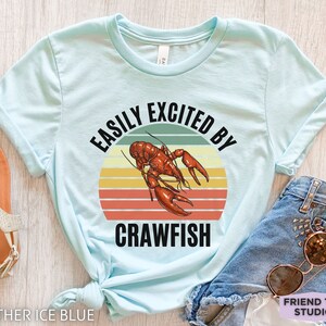 Op de afbeelding: Een lichtblauw T-shirt met een retro zonsondergangafbeelding en een kreeftenillustratie. De tekst "Easily Excited By Crawfish" staat op het shirt gedrukt.