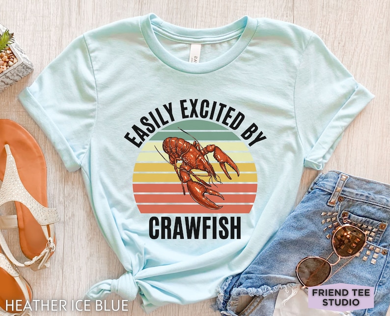 Op de afbeelding: Een lichtblauw T-shirt met een retro zonsondergangafbeelding en een kreeftenillustratie. De tekst "Easily Excited By Crawfish" staat op het shirt gedrukt.