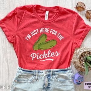 May include: Heather red t-shirt with rolled sleeves. The shirt features the text "I'M JUST HERE FOR THE PICKLES" with a graphic of two green pickles. The shirt is paired with denim shorts and sunglasses.