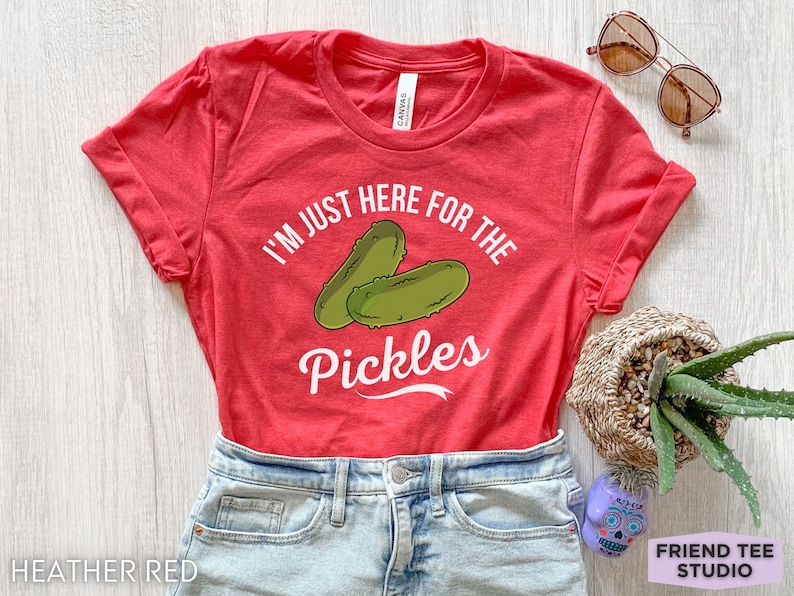 May include: Heather red t-shirt with rolled sleeves. The shirt features the text "I'M JUST HERE FOR THE PICKLES" with a graphic of two green pickles. The shirt is paired with denim shorts and sunglasses.