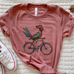 K&ouml;nnte beinhalten: Ein staubrosa T-Shirt mit einem bunten Vogel, der Fahrrad f&auml;hrt. Der Vogel hat einen roten Kamm, blaue und gr&uuml;ne Fl&uuml;gel und sitzt auf einem schwarzen Fahrrad mit goldfarbenen R&auml;dern. Das Shirt hat einen Rundhalsausschnitt und kurze &Auml;rmel.