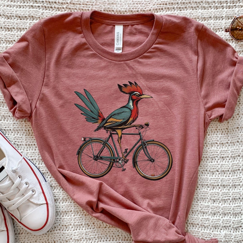 K&ouml;nnte beinhalten: Ein staubrosa T-Shirt mit einem bunten Vogel, der Fahrrad f&auml;hrt. Der Vogel hat einen roten Kamm, blaue und gr&uuml;ne Fl&uuml;gel und sitzt auf einem schwarzen Fahrrad mit goldfarbenen R&auml;dern. Das Shirt hat einen Rundhalsausschnitt und kurze &Auml;rmel.