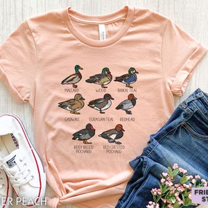 Peut inclure: Un t-shirt &agrave; manches courtes couleur p&ecirc;che avec une impression graphique de huit esp&egrave;ces de canards diff&eacute;rentes, accompagn&eacute;es de leurs noms : Mallard, Wood, Baikal Teal, Gadwall, Eurasian Teal, Redhead, Rosy Billed Pochard et Red Crested Pochard.