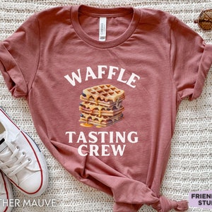May include: Heather mauve t-shirt with the words "WAFFLE TASTING CREW" above a stack of waffles with syrup. The shirt is a crew neck and has rolled-up sleeves. White sneakers are visible on the left side of the image.