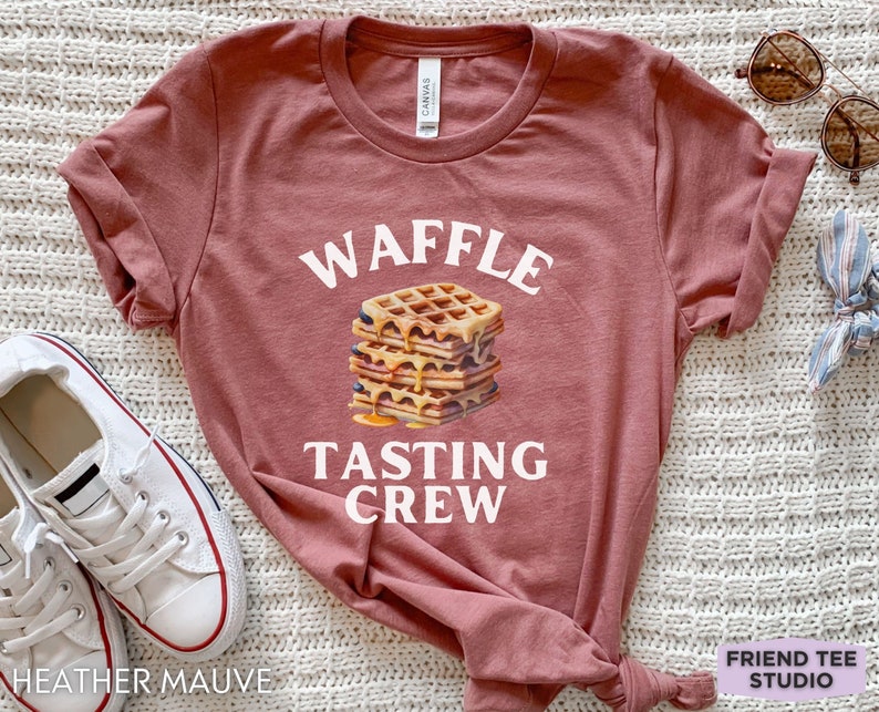 May include: Heather mauve t-shirt with the words "WAFFLE TASTING CREW" above a stack of waffles with syrup. The shirt is a crew neck and has rolled-up sleeves. White sneakers are visible on the left side of the image.