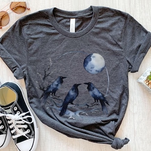 May include: Dark gray t-shirt featuring a watercolor design of three black crows perched on a branch, with a full moon in the background. The shirt has a crew neck and short sleeves. A pair of black and white sneakers and sunglasses are in the image.