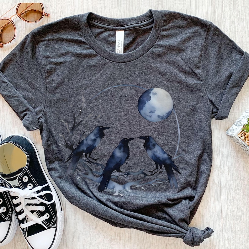 May include: Dark gray t-shirt featuring a watercolor design of three black crows perched on a branch, with a full moon in the background. The shirt has a crew neck and short sleeves. A pair of black and white sneakers and sunglasses are in the image.