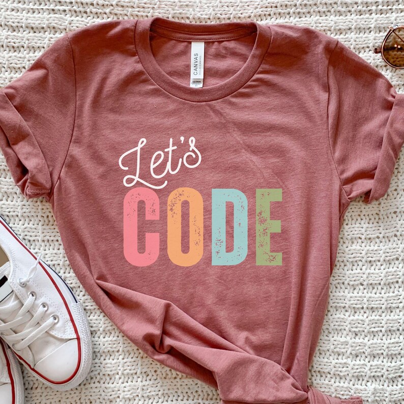 K&ouml;nnte beinhalten: Ein staubrosa T-Shirt mit dem Schriftzug "Let's CODE" in einer farbenfrohen, verspielten Schriftart. Das Wort "Let's" ist in wei&szlig;er Schrift, w&auml;hrend "CODE" in rosa, orange, blau und gr&uuml;nen Blockbuchstaben gehalten ist. Wei&szlig;e Turnschuhe sind unten links zu sehen.