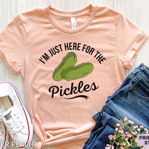 May include: A peach-colored t-shirt with the text "I'M JUST HERE FOR THE PICKLES" and a graphic of two green pickles. The shirt is paired with blue jeans and white sneakers.