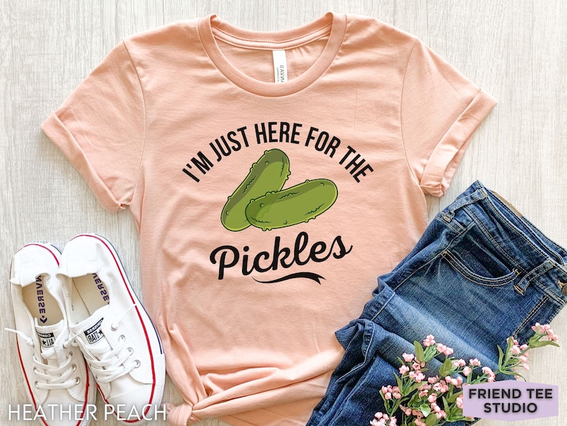 May include: A peach-colored t-shirt with the text "I'M JUST HERE FOR THE PICKLES" and a graphic of two green pickles. The shirt is paired with blue jeans and white sneakers.