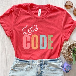 K&ouml;nnte beinhalten: Ein rotes T-Shirt mit den Worten "Let's CODE" in wei&szlig;en und bunten Blockbuchstaben. Das Shirt ist gefaltet, und das Bild enth&auml;lt eine Sonnenbrille und einen dekorativen Korb.