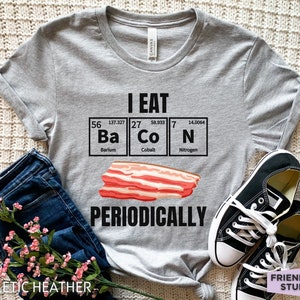 Puede incluir: Camiseta gris jaspeado con el texto "I EAT Ba Co N PERIODICALLY" en negro, con una ilustraci&oacute;n de bacon y elementos de la tabla peri&oacute;dica. La camiseta se combina con vaqueros y zapatillas negras.