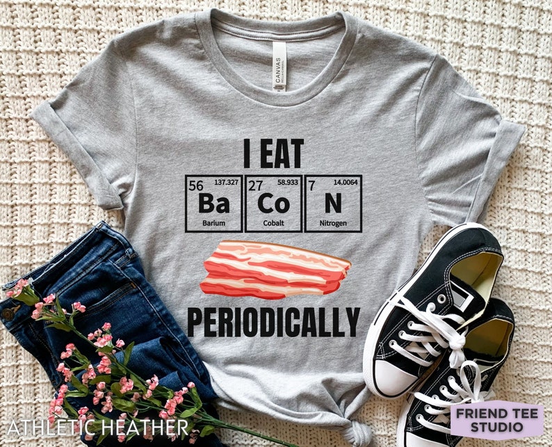 Puede incluir: Camiseta gris jaspeado con el texto "I EAT Ba Co N PERIODICALLY" en negro, con una ilustraci&oacute;n de bacon y elementos de la tabla peri&oacute;dica. La camiseta se combina con vaqueros y zapatillas negras.