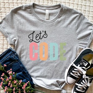K&ouml;nnte beinhalten: Ein meliertes graues T-Shirt mit dem Schriftzug "Let's CODE" in einer farbenfrohen, verspielten Schriftart. Das Shirt wird mit einer blauen Jeans, schwarzen Turnschuhen und einem Zweig rosa Bl&uuml;ten kombiniert.