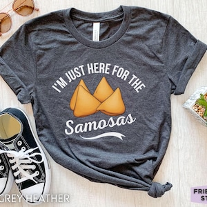 Puede incluir: Una camiseta de manga corta en gris jaspeado oscuro con un gráfico de tres samosas doradas en el centro. El texto en blanco sobre y bajo el gráfico dice: I'M JUST HERE FOR THE Samosas. La camiseta se presenta junto a zapatillas negras y blancas.