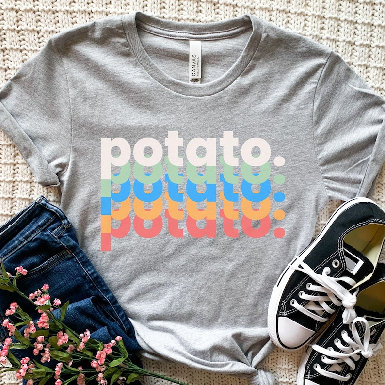 May include: A heather gray short-sleeve t-shirt featuring the word potato printed in four stacked rows of varying colors including light pink, light green, blue, and orange, with a period at the end of each row, paired with dark blue denim jeans.
