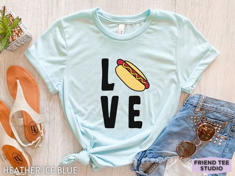May include: A light blue t-shirt with the word "LOVE" in black letters. The "O" is replaced with a cartoon hot dog with mustard and ketchup.