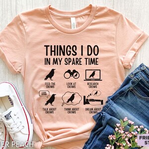 K&ouml;nnte beinhalten: Ein pfirsichfarbenes T-Shirt mit schwarzem Text, der "Things I do in my spare time" lautet. Der Text ist umgeben von schwarzen Kr&auml;henillustrationen und Symbolen, die Aktivit&auml;ten im Zusammenhang mit Kr&auml;hen darstellen, wie z. B. das F&uuml;ttern, Betrachten, Recherchieren, Reden, Denken und Tr&auml;umen von Kr&auml;hen.