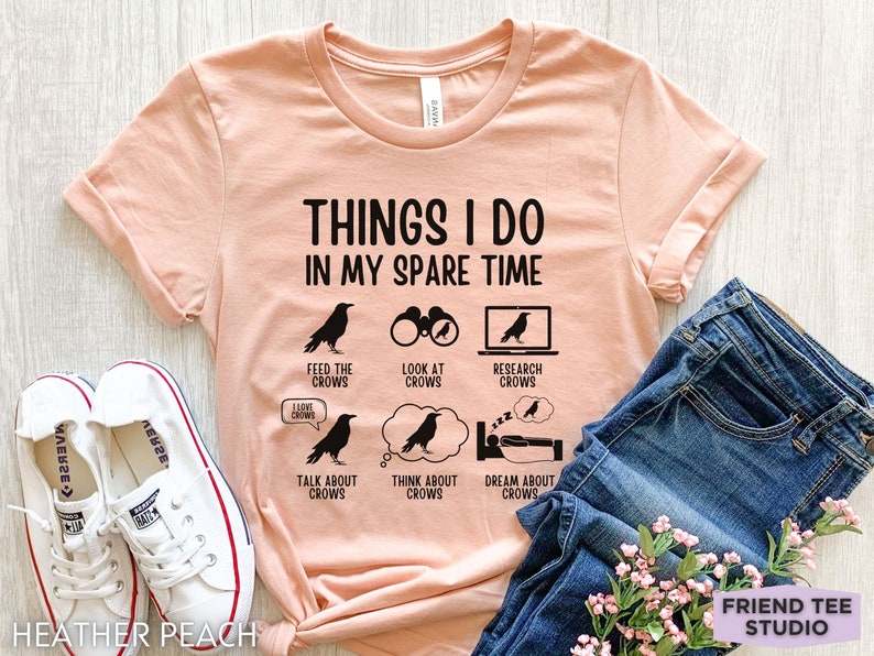 K&ouml;nnte beinhalten: Ein pfirsichfarbenes T-Shirt mit schwarzem Text, der "Things I do in my spare time" lautet. Der Text ist umgeben von schwarzen Kr&auml;henillustrationen und Symbolen, die Aktivit&auml;ten im Zusammenhang mit Kr&auml;hen darstellen, wie z. B. das F&uuml;ttern, Betrachten, Recherchieren, Reden, Denken und Tr&auml;umen von Kr&auml;hen.