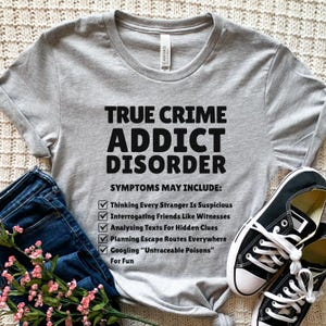 Puede incluir: Una camiseta gris claro con el texto negro "TRUE CRIME ADDICT DISORDER". La camiseta enumera síntomas que incluyen "Pensar que todos los extraños son sospechosos" y "Buscar en Google 'Veneno indetectable'". También se ven zapatillas de lona negras y vaqueros azules.