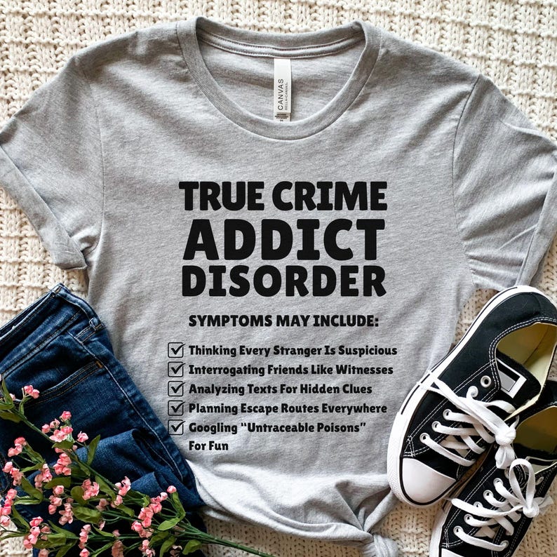 May include: A grey t-shirt with the text "TRUE CRIME ADDICT DISORDER" in black. The shirt lists symptoms including "Thinking Every Stranger Is Suspicious" and "Googling 'Untraceable Poisons'". Black canvas trainers and blue jeans are also visible.
