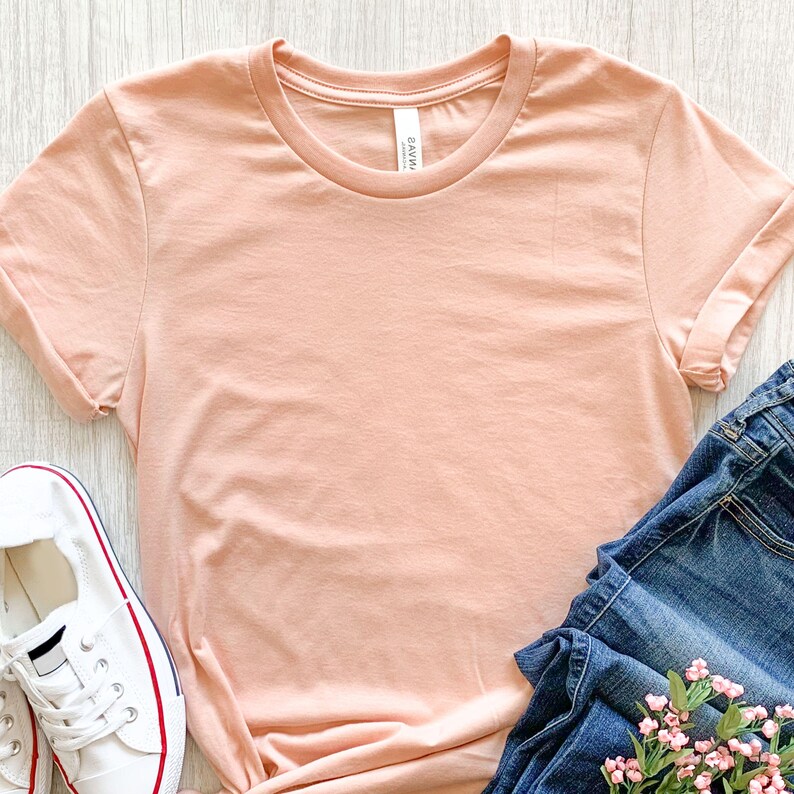 May include: A peach colored t-shirt with a crew neck and rolled up sleeves. The t-shirt is laying flat on a wooden surface with a pair of blue jeans and a bouquet of pink flowers.