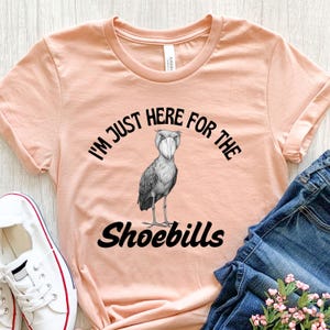 May include: A peach-colored t-shirt with a black graphic of a shoebill stork wearing a face mask. The text "I'm just here for the shoebills" is printed on the shirt.