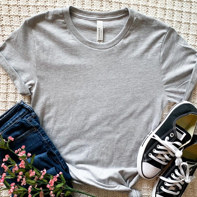 May include: A heather gray t-shirt with a white tag that says "Canvas" on it. The t-shirt is laying on a white fuzzy surface with a pair of black canvas shoes and blue jeans with pink flowers.