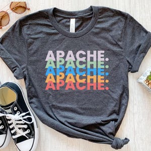 May include: Dark gray t-shirt with the word "APACHE" stacked in multiple colors: lavender, light green, blue, orange, and red. The shirt is made of a soft material and has a crew neck.