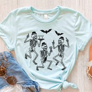 May include: A light blue t-shirt featuring a black graphic of dancing skeletons wearing bowler hats and holding drinks. Two bats are above the skeletons. The shirt is tied at the waist.