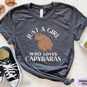 May include: Dark grey heather t-shirt with white text that reads "Just a girl who loves capybaras". A cartoon capybara is pictured above the text.