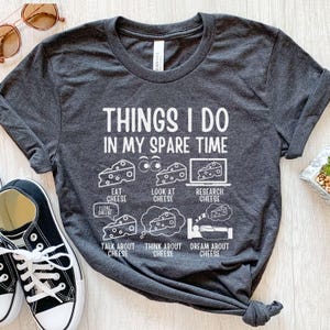 Puede incluir: Una camiseta gris oscuro con un gr&aacute;fico blanco que dice "Cosas que hago en mi tiempo libre" y enumera actividades relacionadas con el queso. Las actividades incluyen comer queso, mirar queso, investigar queso, hablar de queso, pensar en queso y so&ntilde;ar con queso.