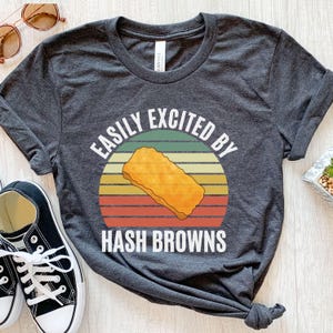 May include: Charcoal grey t-shirt with the words "EASILY EXCITED BY HASH BROWNS" in white. The design features a retro-style graphic of a hash brown on a circular background with orange, yellow, green, and red horizontal stripes.