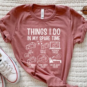 Puede incluir: Una camiseta color marr&oacute;n con un gr&aacute;fico blanco que dice "Cosas que hago en mi tiempo libre" y presenta una serie de actividades relacionadas con el queso, incluyendo comer, mirar, investigar, hablar, pensar y so&ntilde;ar con queso.