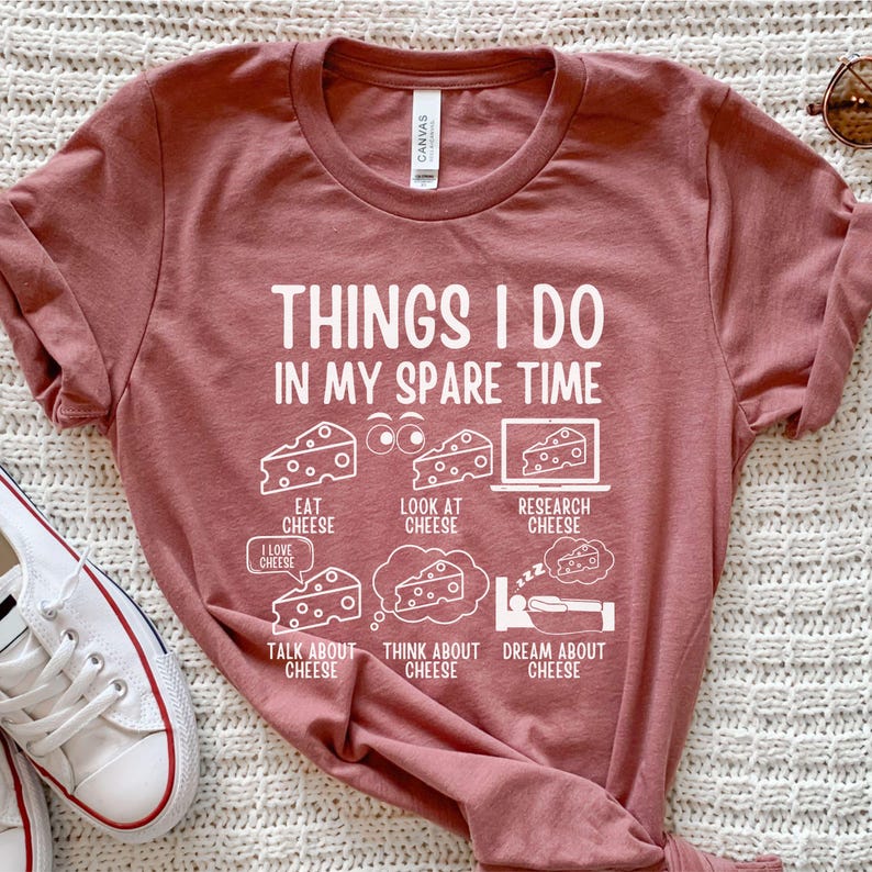 Puede incluir: Una camiseta color marr&oacute;n con un gr&aacute;fico blanco que dice "Cosas que hago en mi tiempo libre" y presenta una serie de actividades relacionadas con el queso, incluyendo comer, mirar, investigar, hablar, pensar y so&ntilde;ar con queso.