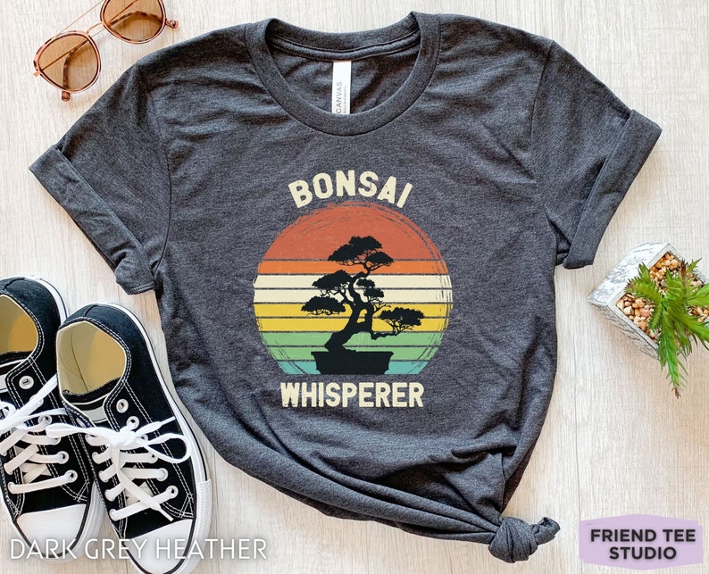 May include: Dark grey heather t-shirt with a vintage-style sunset graphic and the text "Bonsai Whisperer".