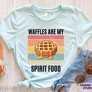 Pu&ograve; includere: T-shirt azzurra con la scritta "WAFFLES ARE MY SPIRIT FOOD" sopra una grafica di waffle con sciroppo. Il design presenta una combinazione di colori retr&ograve; arancione, giallo e blu. La maglietta &egrave; realizzata in materiale morbido.