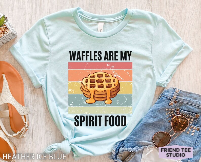 Pu&ograve; includere: T-shirt azzurra con la scritta "WAFFLES ARE MY SPIRIT FOOD" sopra una grafica di waffle con sciroppo. Il design presenta una combinazione di colori retr&ograve; arancione, giallo e blu. La maglietta &egrave; realizzata in materiale morbido.