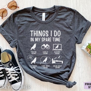 K&ouml;nnte beinhalten: Dunkelgraues T-Shirt aus Heather-Material mit wei&szlig;em Text, der "Things I do in my spare time" lautet. Der Text ist von Illustrationen von Raben in verschiedenen Aktivit&auml;ten umgeben, darunter das F&uuml;ttern, Betrachten, Recherchieren, Reden &uuml;ber, Nachdenken &uuml;ber und Tr&auml;umen von Raben.