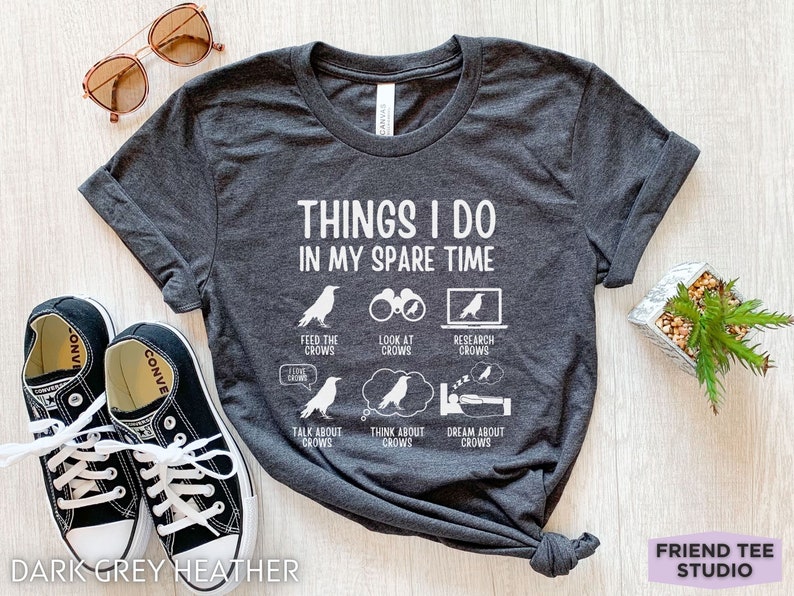 K&ouml;nnte beinhalten: Dunkelgraues T-Shirt aus Heather-Material mit wei&szlig;em Text, der "Things I do in my spare time" lautet. Der Text ist von Illustrationen von Raben in verschiedenen Aktivit&auml;ten umgeben, darunter das F&uuml;ttern, Betrachten, Recherchieren, Reden &uuml;ber, Nachdenken &uuml;ber und Tr&auml;umen von Raben.